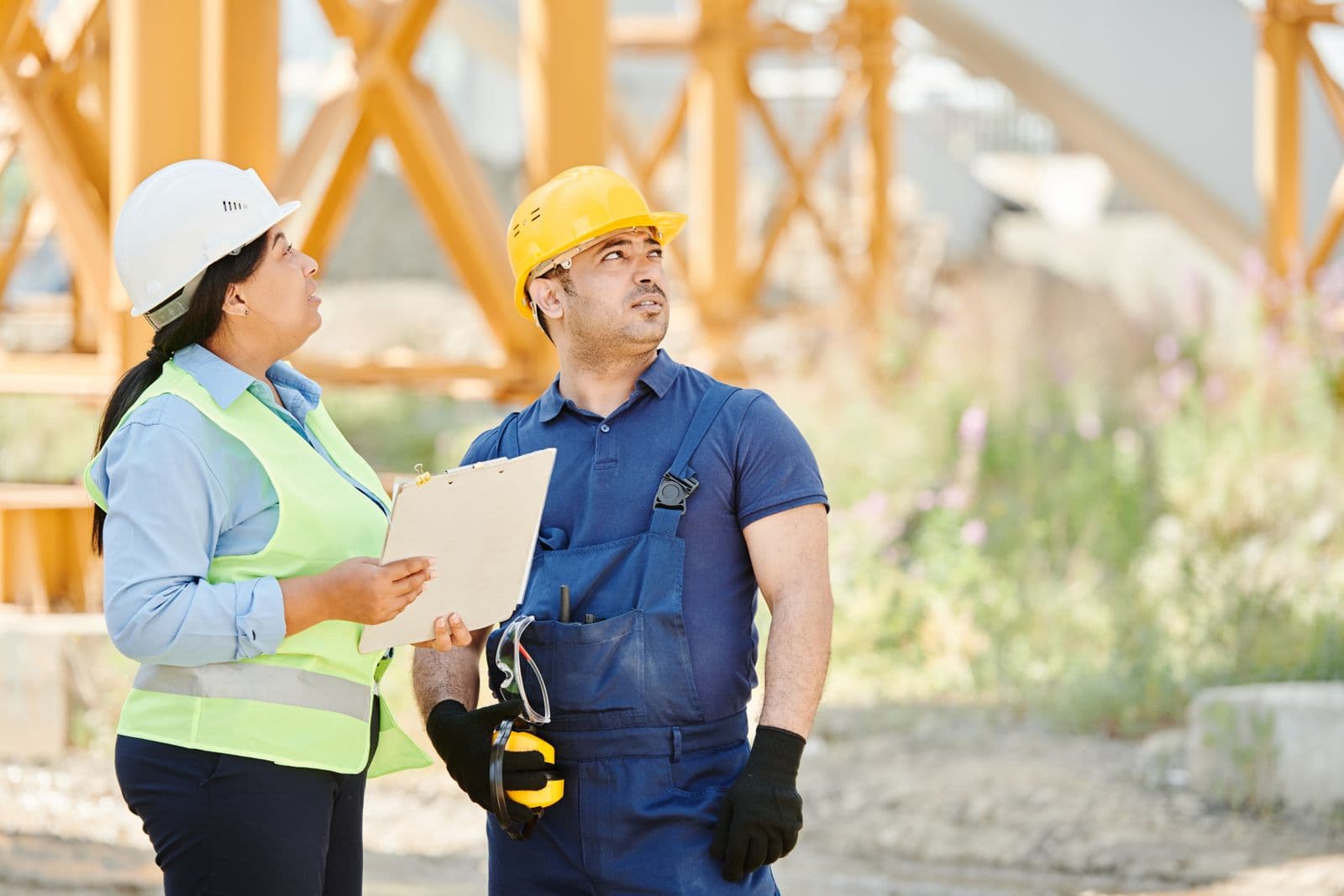 Two workers in hard hats and hi-vis vests reviewing site plans on a clipboard outdoors.